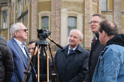 Dudás Tibor kancellár középen áll, előtte egy nagy videó állvány, körülötte kormány emberei, pl. Rétvári Bence, mögötte a Király Endre téglaépülete