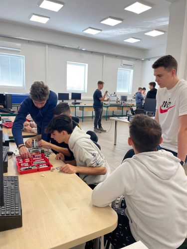 Diákok  (fiúk, öten ) a DKA (digitális közösségi alkotótér) teremben egy számítógép előtt ülnek és apró lego robot építőelmeket választanak ki a egy műanyag rekeszből.
