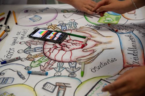 Kézzel készült színes rajz az asztalon, ami egy szívet tartó kezet ábrázol, fölötte a "közös ügyünk az ápolás"-szöveggel és a rajzoló kezei is a képen vannak