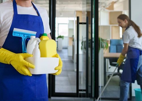 Egy férfi takarító előtérben munkaruhában tart egy dobozt, benne takarítóeszközökkel, a háttérben egy női takarító takarít egy partvissal.