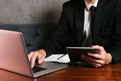 Öltönyös üzletember ül egy íróasztalnál, jobb kezével egy laptopon gépel, bal kezében egy tablet van